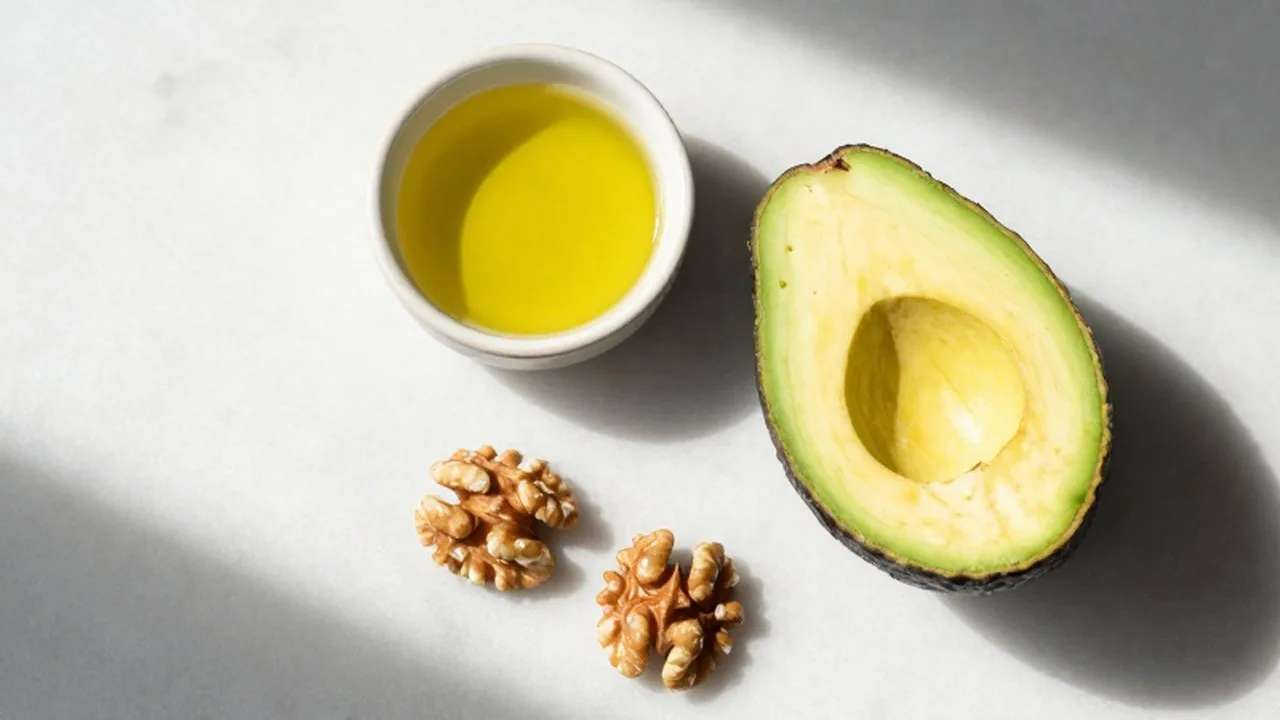 Aguacate partido a la mitad, nueces y un pequeño cuenco de aceite de oliva sobre mármol blanco.