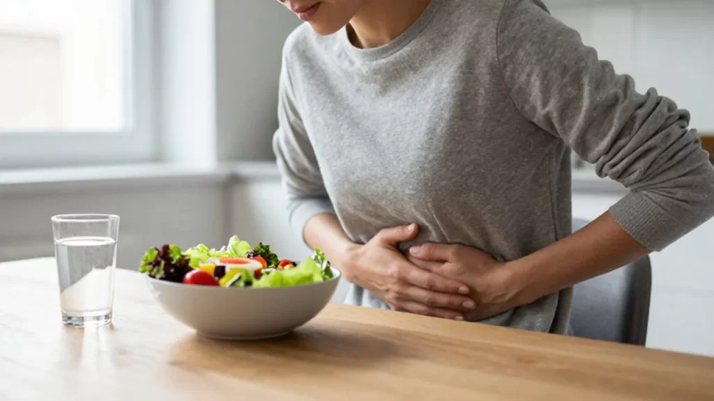 Persona sentada a la mesa de una cocina con una ensalada y tocándose el abdomen.